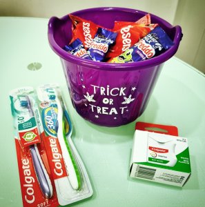 Image of a purple Trick or Treat Bucket filled with chocolate, surrounded by two toothbrushes and a floss 